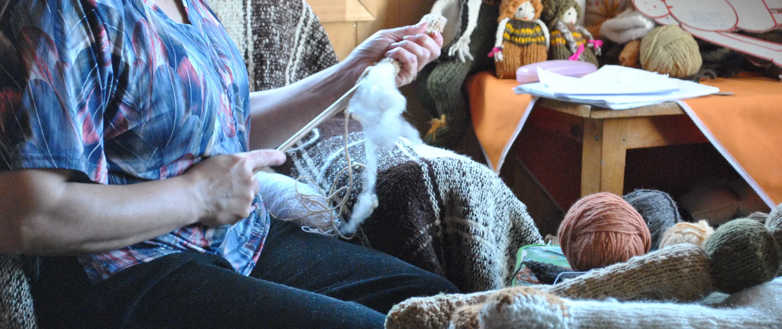 Persona sentada rellena con vellón blanco una pierna de muñeca tejida, rodeada de ovillos de lana de colores y tejidos de lana en un espacio interior.