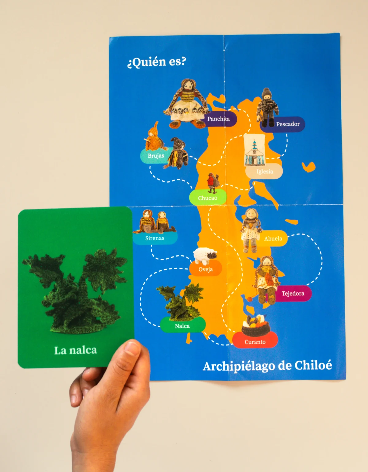 Mano sostiene una tarjeta de color verde con la imagen de una nalca tejida, sobre un mapa del archipiélago de Chiloé.