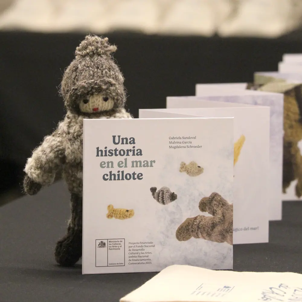Muñeco tejido de lana con gorro y ropa gris junto al libro Una historia en el mar chilote, que muestra figuras de peces en su portada.