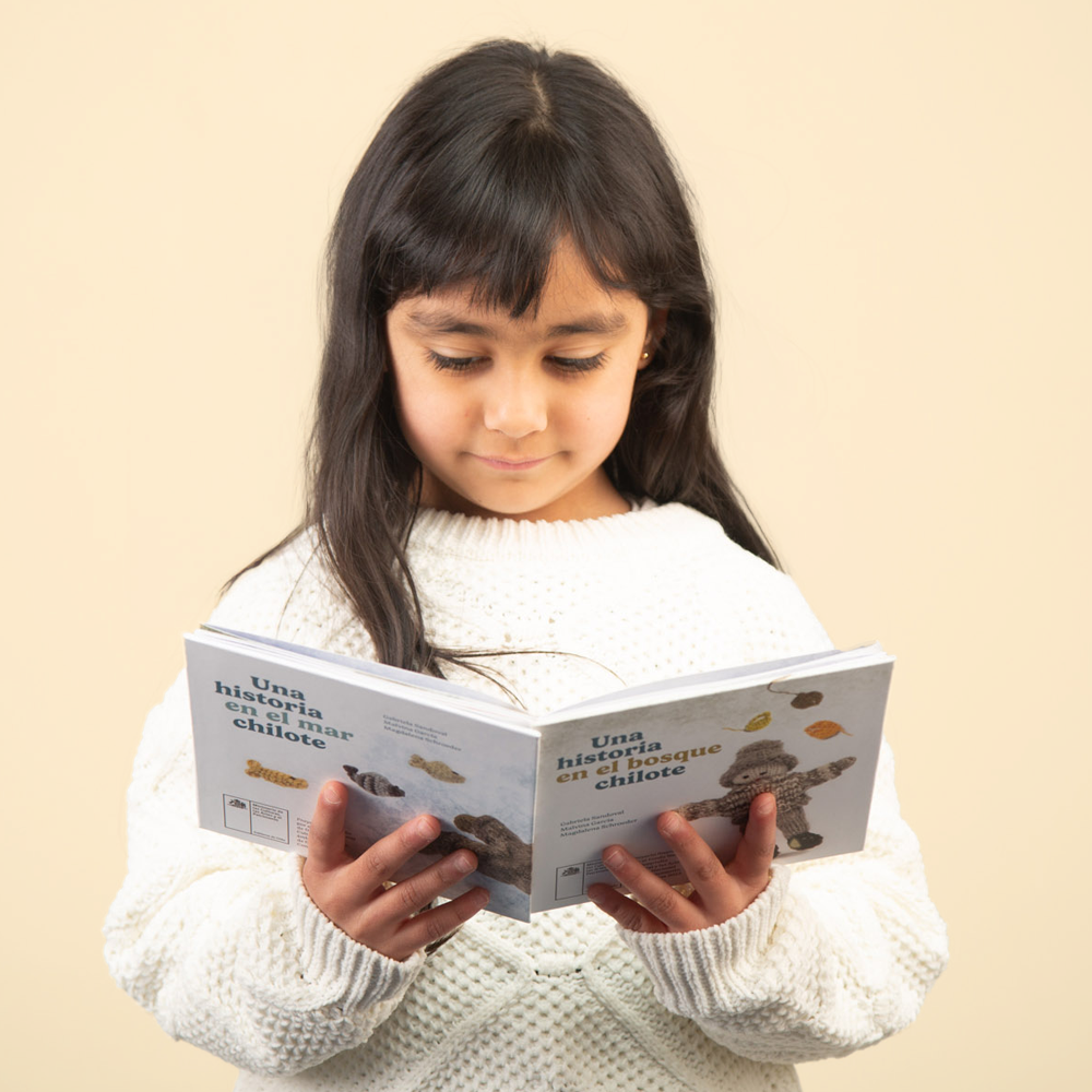 Niña con suéter blanco sostiene con dos manos el libro del pescador de chiloé y lo lee.