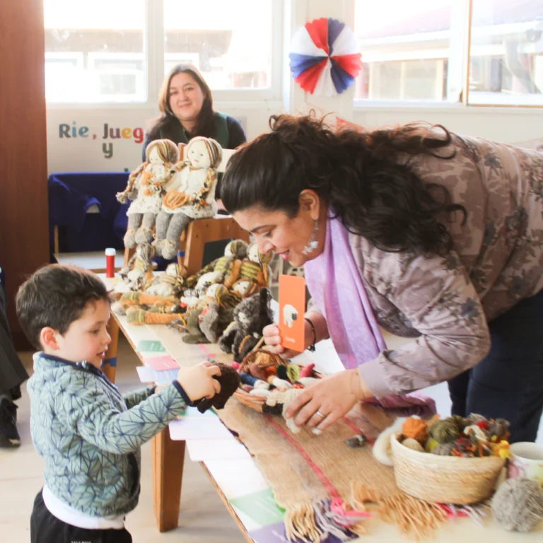 Mujer muestra muñecos de lana a un niño pequeño en una mesa con tejidos y materiales artesanales. Al fondo hay otra mujer y decoración con cintas rojas, blancas y azules.