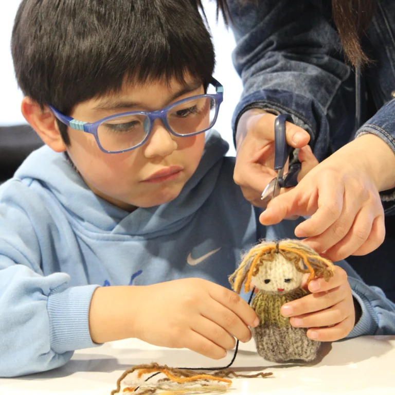 Niño sostiene un muñeco tejido y un trozo de lana, mientras unas manos adultas le ayudan a cortarlo.