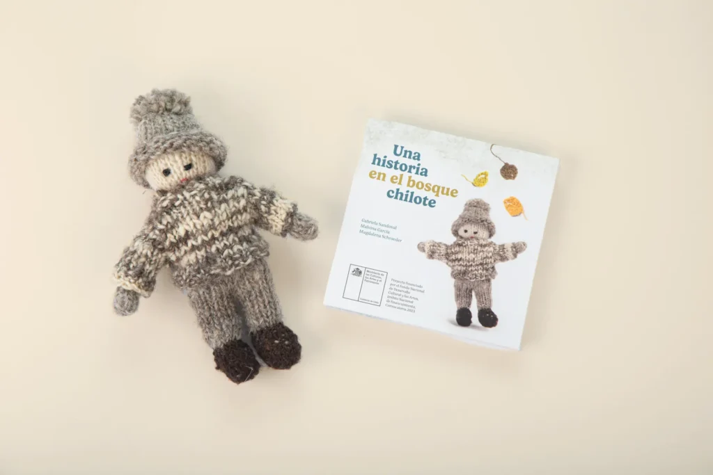 Muñeco tejido de lana con gorro y ropa gris junto al libro Una historia en el bosque chilote, que muestra la misma figura en su portada.