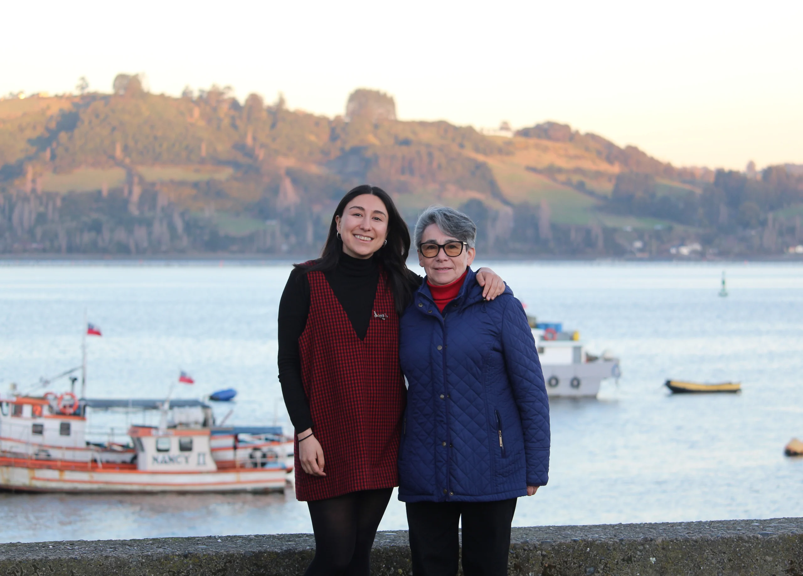 La diseñadora Gabriela Sandoval y la artesana Malvina García se abrazan frente a las costas de Chiloé y un cielo despejado.
