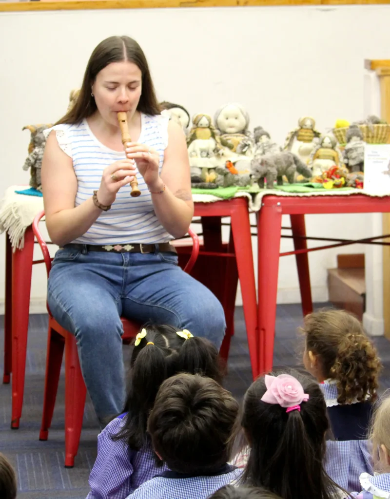 Mediadora toca una flauta dulce frente a un grupo de niños y niñas sentados en el suelo.