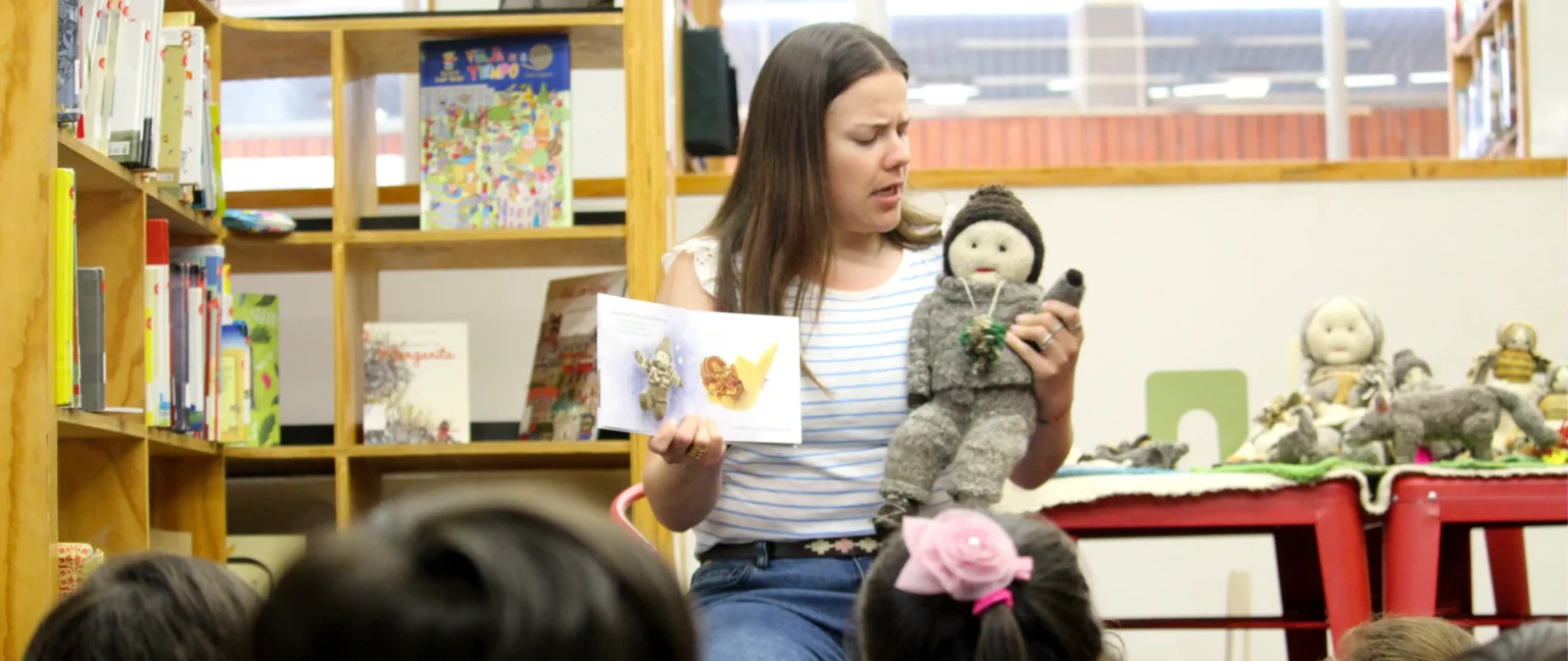 Mediadora sostiene un libro ilustrado desplegable y un muñeco tejido mostrándolo a un grupo de niños y niñas.