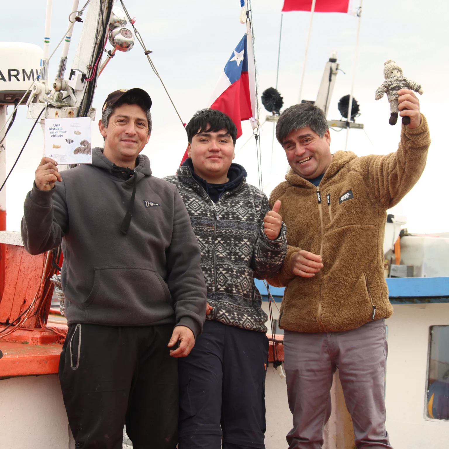 Tres hombres posan en una embarcación. Uno sostiene el libro Una historia en el bosque chilote y otro muestra un muñeco tejido, con banderas chilenas al fondo.