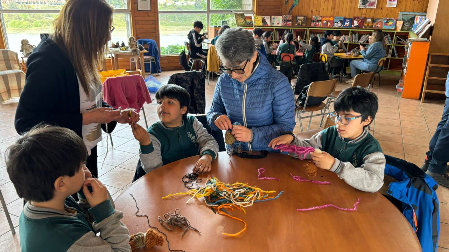 Niños participan en un taller de personalizar personajes de lana dentro de la biblioteca. Están sentados alrededor de una mesa de madera, manipulando hilos de colores y pequeños tejidos de lana. Dos mujeres los acompañan y orientan mientras al fondo se ven estanterías con libros y otras personas trabajando.