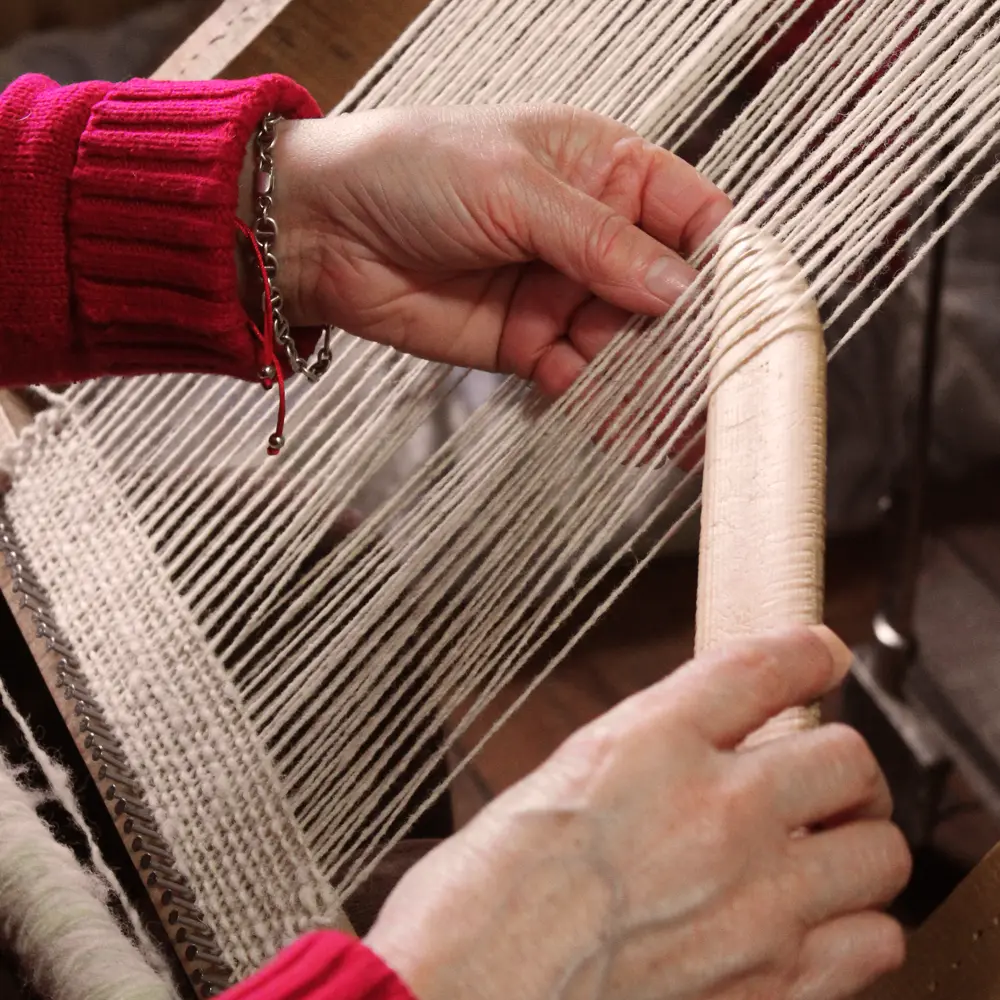 Manos de una persona tejiendo con lana clara en un telar de madera, moviendo una lanzadera entre los hilos.