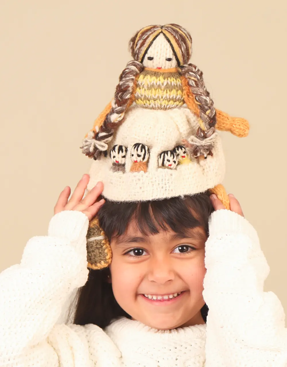 Niña sonriente con suéter blanco sostiene sobre su cabeza una muñeca tejida de lana llamada Panchita. La muñeca tiene trenzas largas y un vestido beige que con cinco bolsillos donde guarda a sus cinco hijos.
