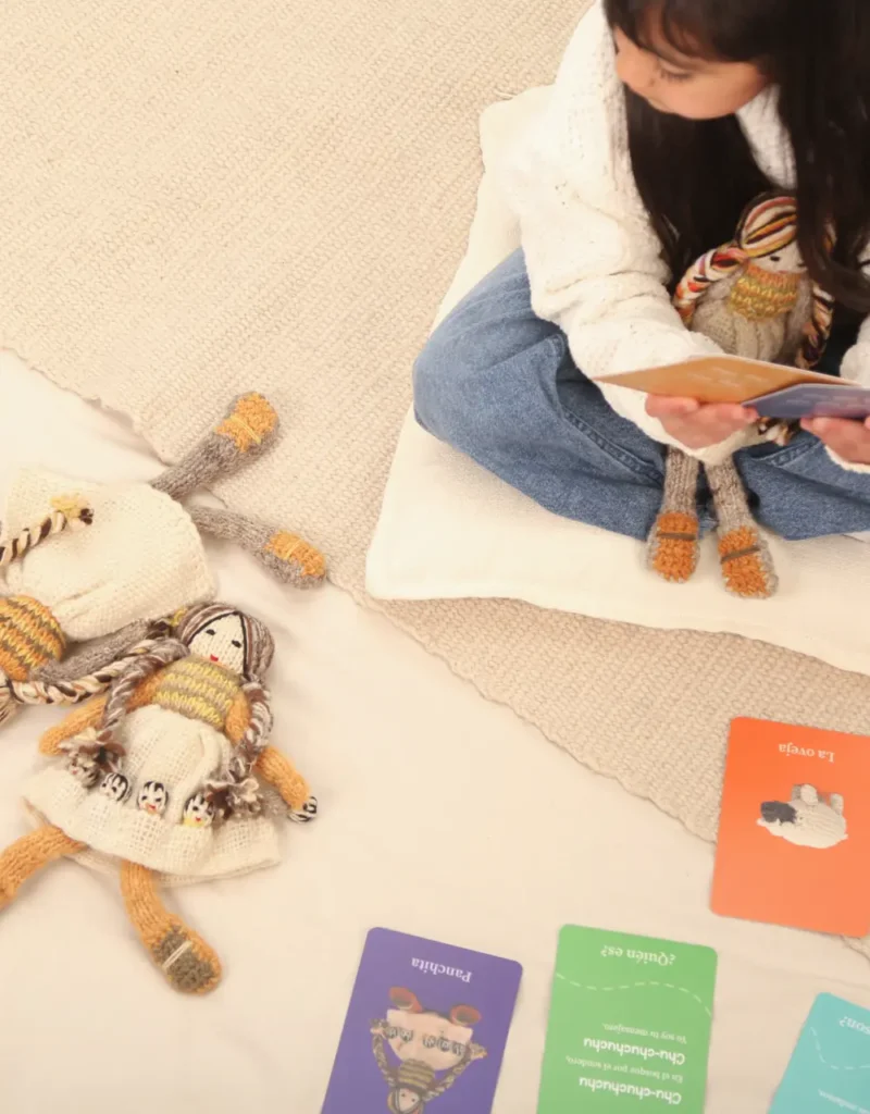 Niña sentada sobre un cojín sostiene una muñeca tejida y una tarjeta del set Panchita recorre Chiloé. En el suelo hay otra muñeca y varias tarjetas de colores.