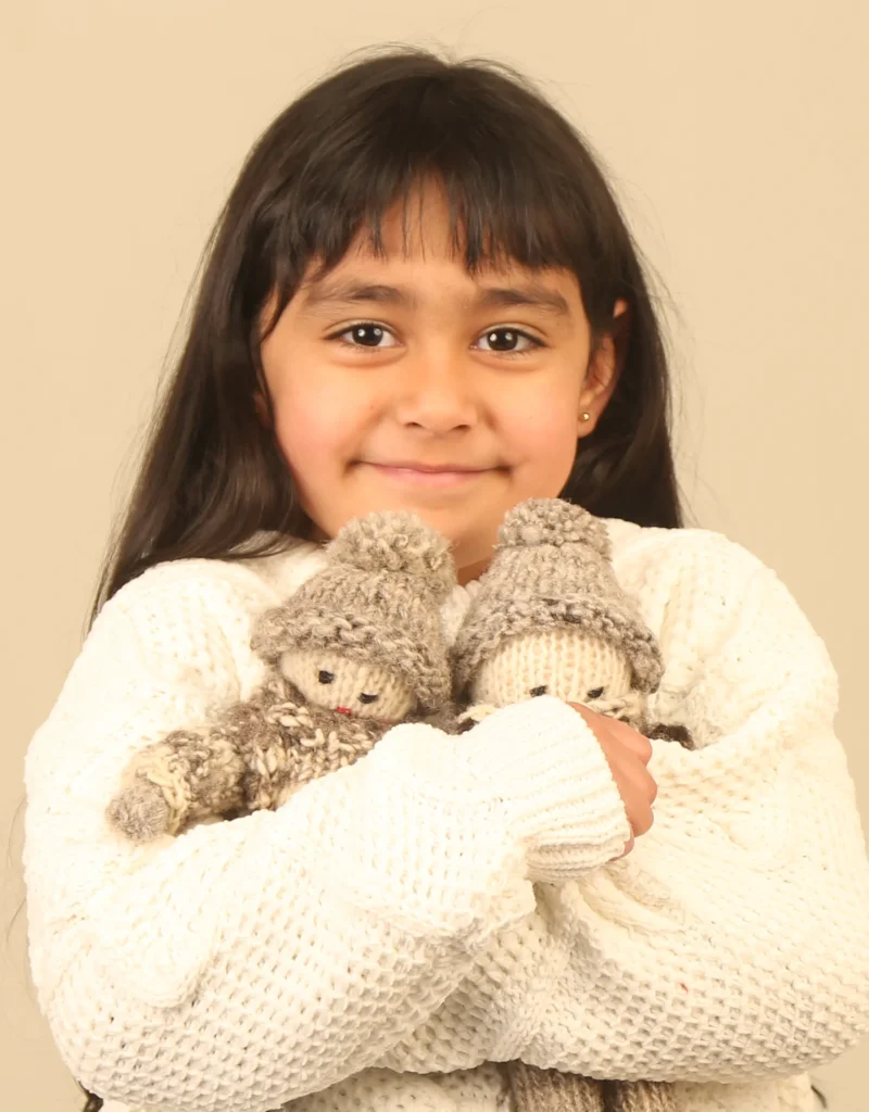 Niña sonriente con suéter blanco abraza a dos muñecos tejidos de lana. Estos muñecos representan al pescador de Chiloé y usan un grueso chaleco tejido y gorro de color gris.