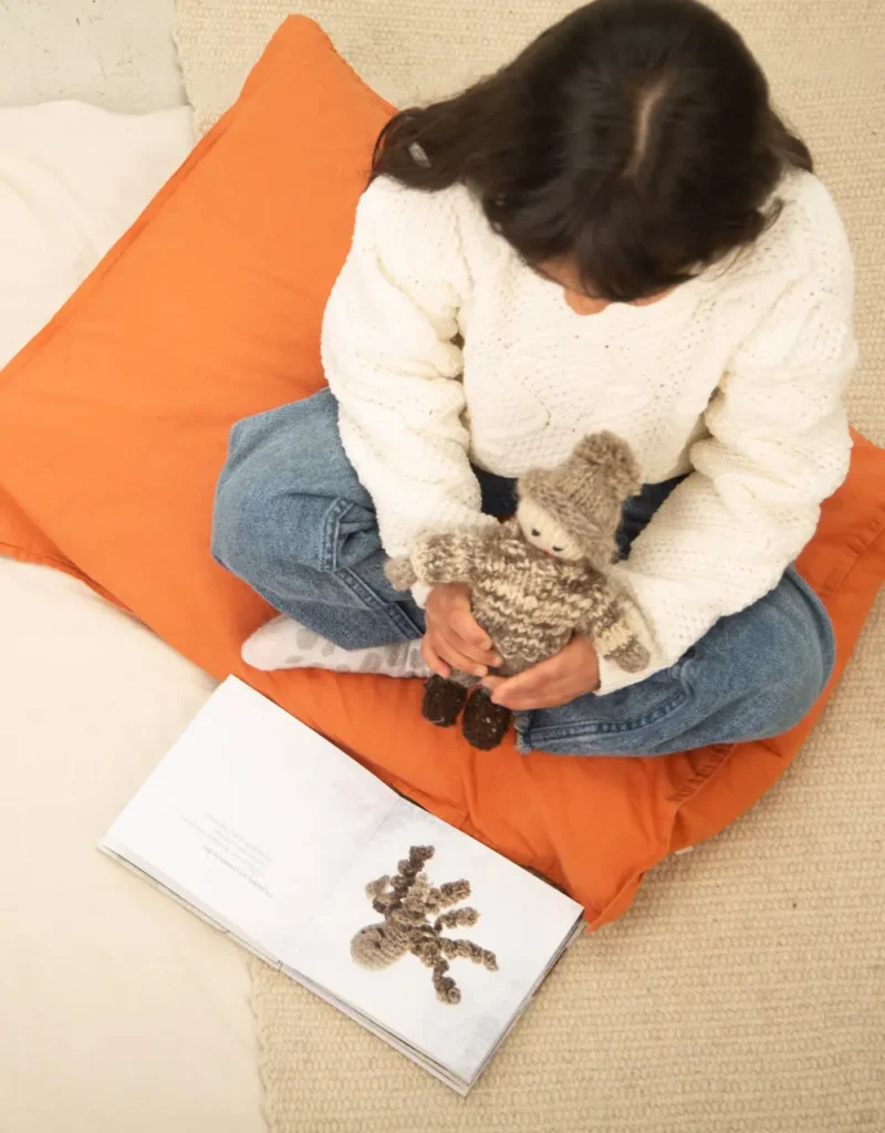 Niña sentada sobre un cojín sostiene un muñeco tejido del pescador de Chiloé mientras lee un libro que está sobre el suelo.