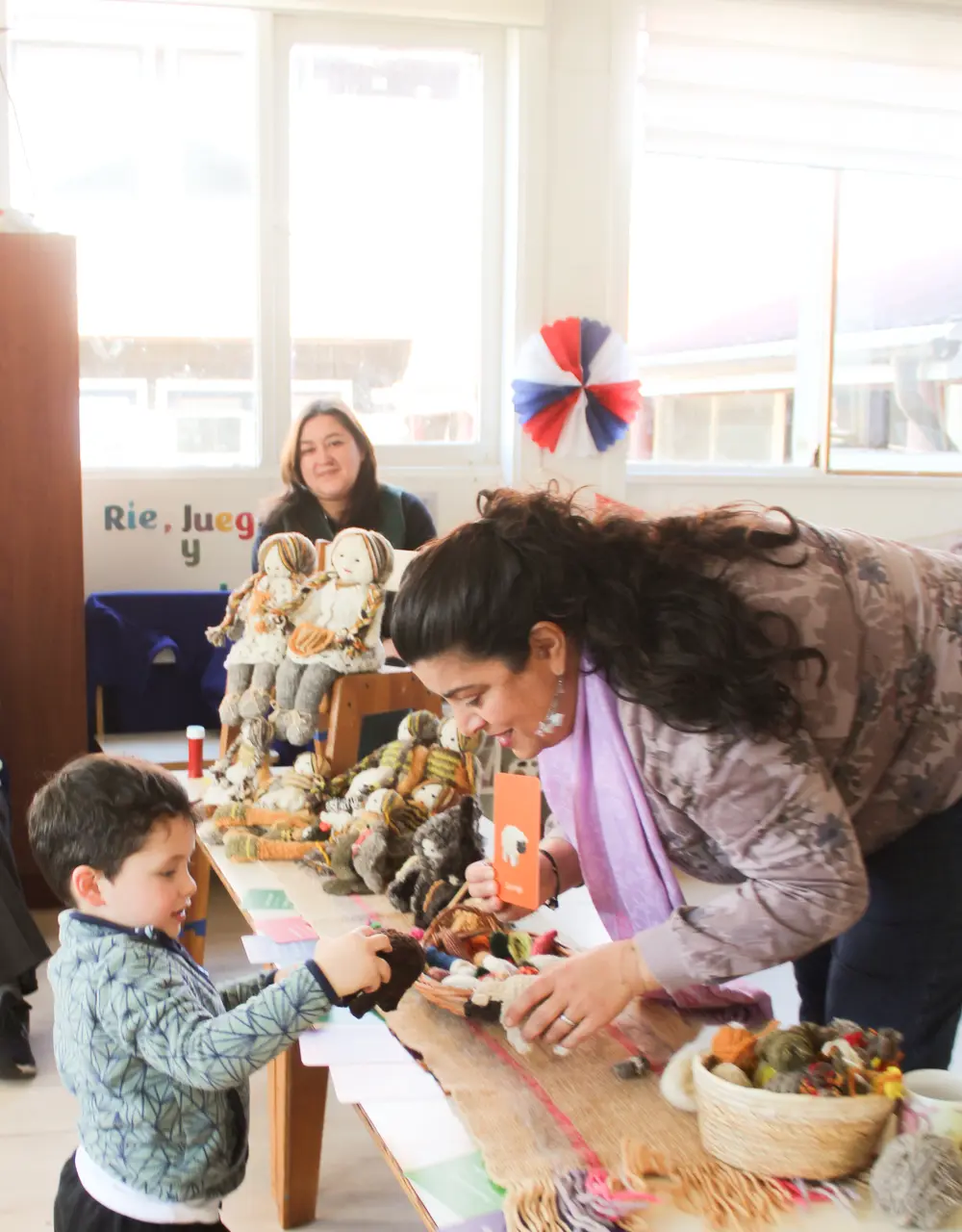 Mujer muestra muñecos de lana a un niño pequeño en una mesa con tejidos y materiales artesanales. Al fondo hay otra mujer y decoración con cintas rojas, blancas y azules.